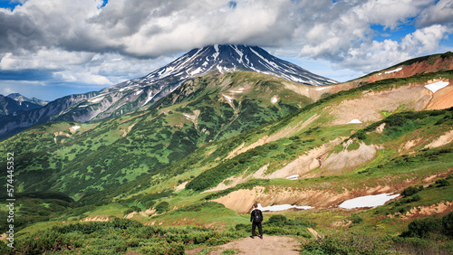 The traveler stands against the backdrop of the Vilyuchensky large volcano, mountains, green landscape, clouds, sunny day. Kamchatka.