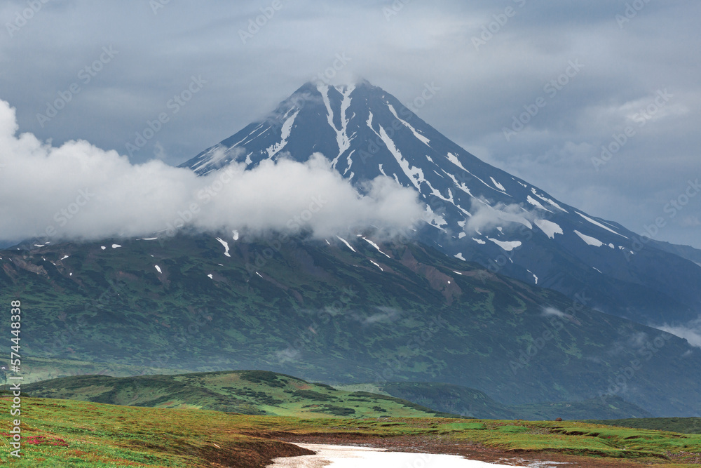 Vilyuchensky volcano on a cloudy day, mountains around, green meadows, snow lies, clouds.