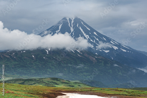 Vilyuchensky volcano on a cloudy day, mountains around, green meadows, snow lies, clouds.