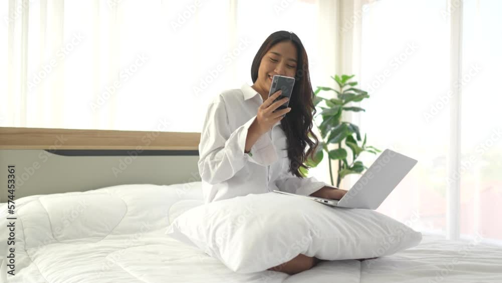 young asian woman using smartphone and laptop when sitting on a bed ...
