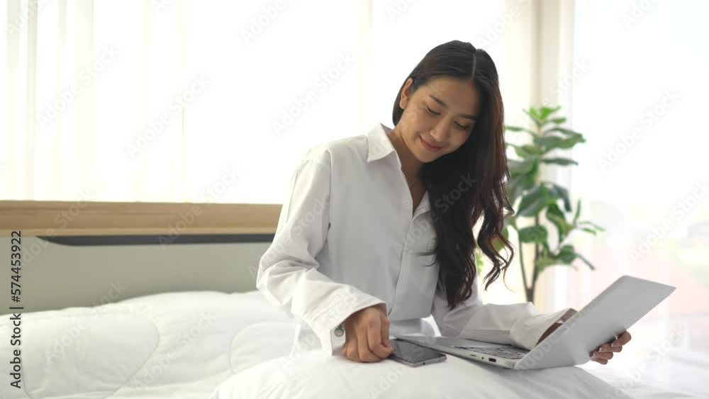 young asian woman using smartphone and laptop when sitting on a bed ...