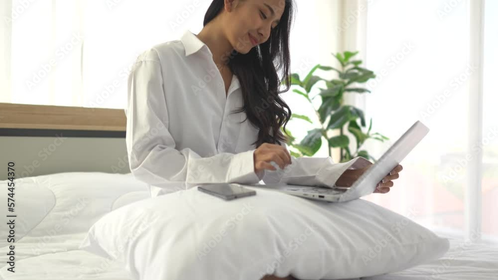 young asian woman using smartphone and laptop when sitting on a bed ...