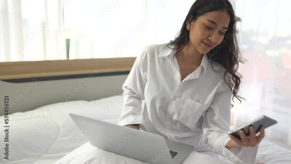young asian woman using smartphone and laptop when sitting on a bed ...