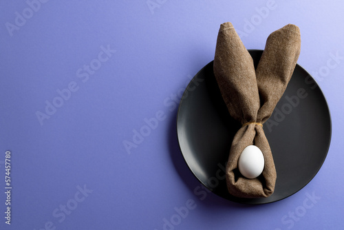 Image of white easter egg and bunny ears on black plate and copy space on purple background