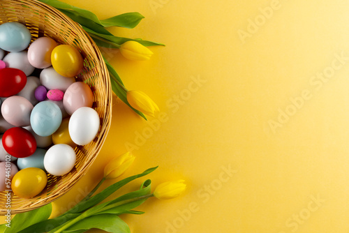 Image of multi coloured easter eggs in basket with yellow tulips and copy space on yellow background
