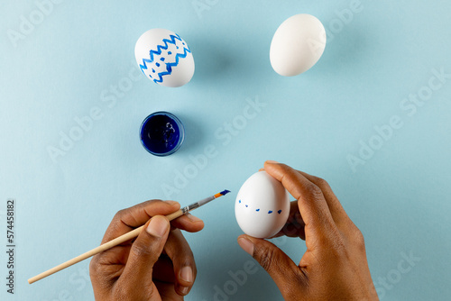 Image of hands of african american woman painting easter eggs with copy space on blue background