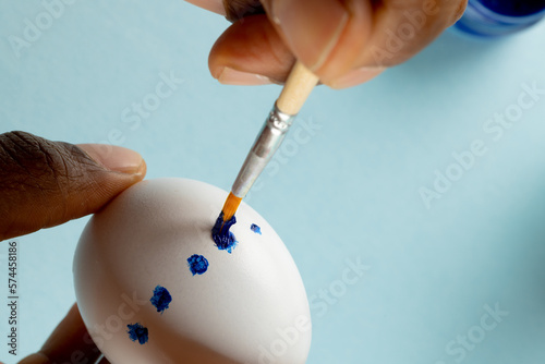 Image of hands of african american woman painting easter egg with copy space on blue background