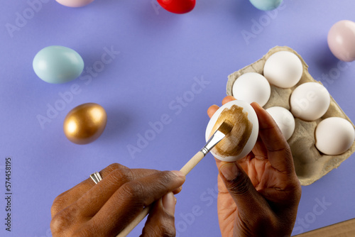 Image of hands of african american woman painting easter eggs with copy space on purple background