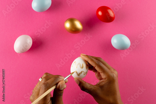 Image of hands of african american woman painting easter eggs with copy space on pink background