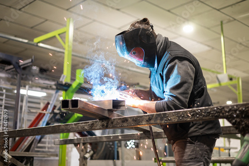 A man working as a welder in a protective mask and work clothes performs work with a MIG-MAG welding machine. Production of welding works.