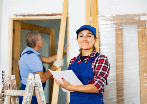 Cheerful Latin female builder in uniform writes remarks in the home construction blueprint