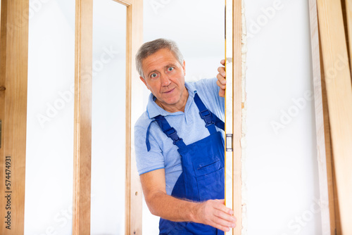 Professional middle-aged repairman in a workwear levelling doorjamb while using waterpass indoors