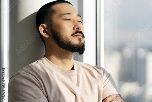 Asian man with closed eyes close-up standing by the window, thinking about something. A young guy takes a break from work