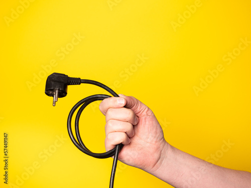 A hand holds a plug to which a black cable is attached.  No face, yellow background.
