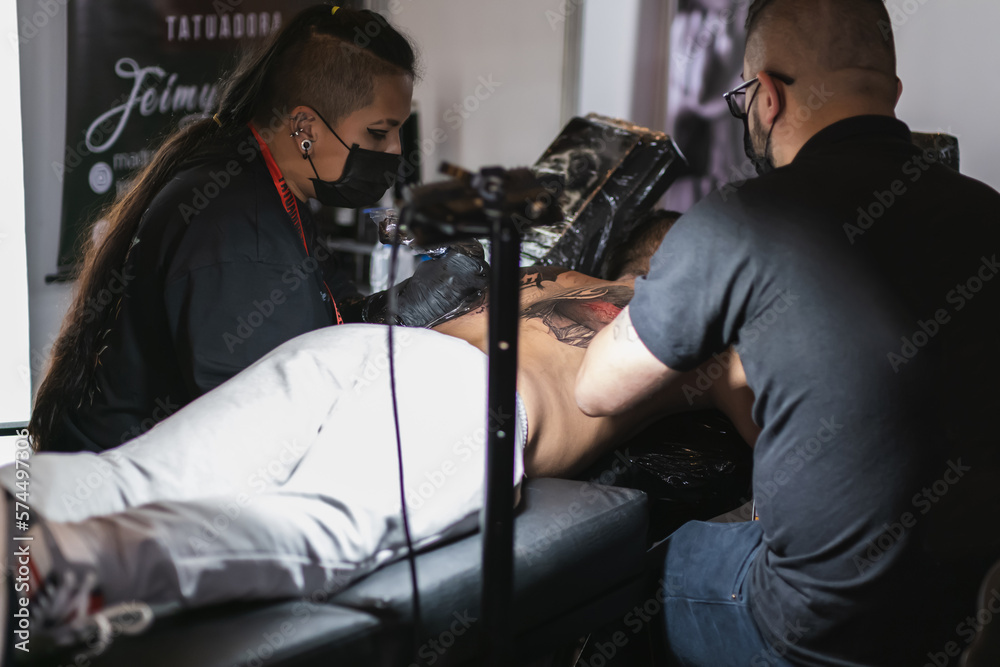 woman and man tattoo artists working at the same time on the back of a man lying down, use of biosecurity, art fusion