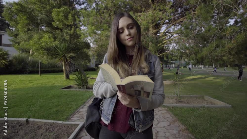 Mujer joven camina por el parque leyendo un libro durante el atardecer.