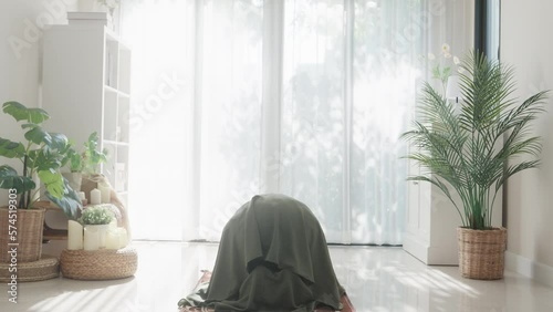 Asian muslim woman sitting on prayer mat pray to allah wear prayer clothes green mukena cover hijab in living room at home, praying room bright light, Islamic faith, Ramadan Kareem, Eid Mubarak. photo