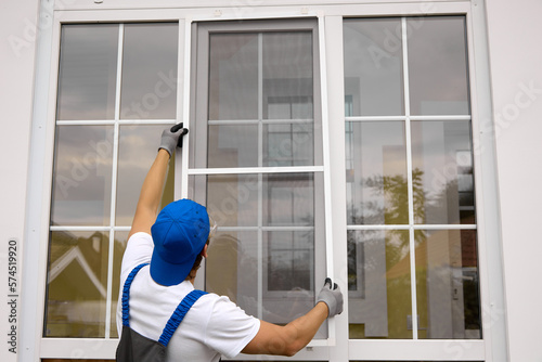 View from back master in working uniform of blue color, in his hands high raises mosquito net for installation on window. Professional installation of protective nets on windows of different sizes