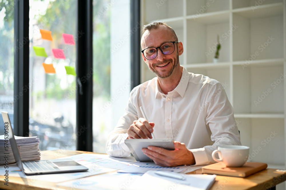 Happy handsome Caucasian businessman sits at his office desk with digital tablet Happy handsome Caucasian businessman sits at his office desk with digital tablet