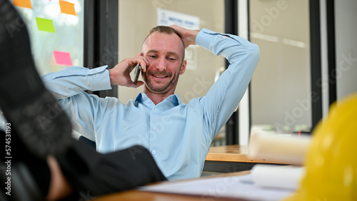 Relaxed Caucasian male engineer putting his legs on table while talking on the phone with client