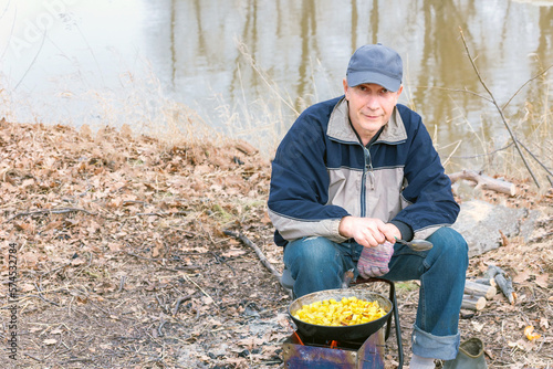 a mature male tourist roasts potatoes in a frying pan on a wood-burning stove in nature on a spring day