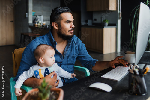 Hispanic father using computer to work at home while he take care of his baby son in Mexico Latin America, home office concept