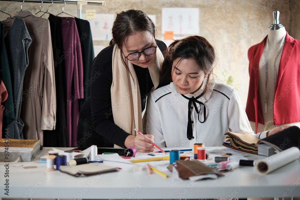 Asian middle-aged female fashion designer teaches a young teen trainee tailor in studio with colorful thread and sewing fabric for dress design collection ideas, professional boutique small business.