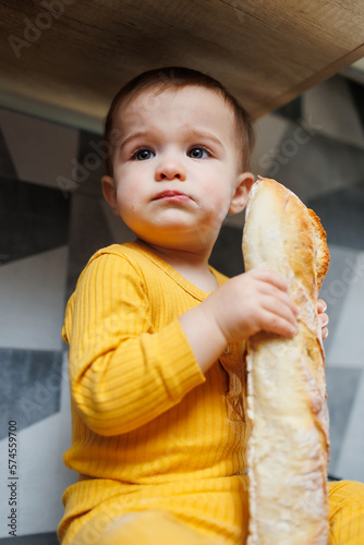 A one-year-old little boy in yellow clothes sits and eats freshly baked rye bread. The child holds a fresh baguette in his hands.