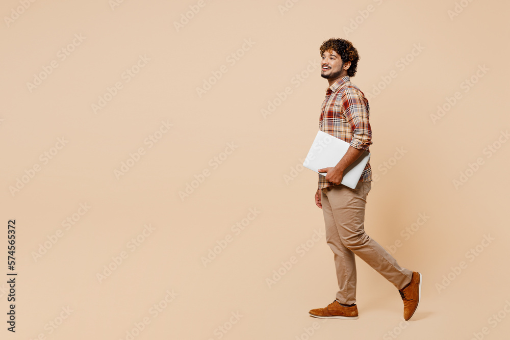 Fototapeta premium Full body side view young IT Indian man wear brown shirt casual clothes hold closed laptop pc computer walk go isolated on plain pastel light beige background studio portrait People lifestyle concept