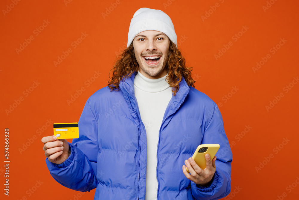 Young man with long curly hair wear hat purple ski padded jacket casual clothes using mobile cell phone hold credit bank card do online shopping booking tour isolated on plain orange red background Young man with long curly hair wear hat purple ski padded jacket casual clothes using mobile cell phone hold credit bank card do online shopping booking tour isolated on plain orange red background