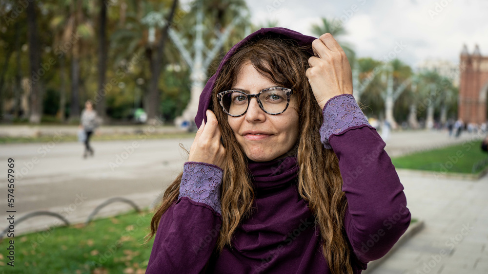Beautiful girl with a purple hood and glasses and curly hair looking into the camera with both hands placed side and side of the face in a park outdoor environment