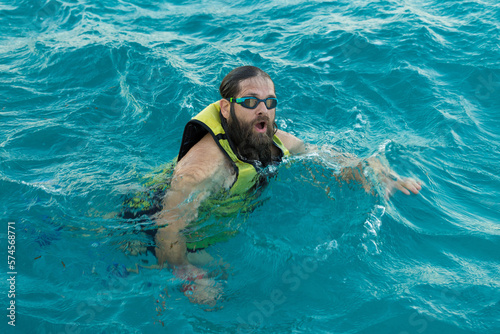 A bearded man diver in a life jacket swims in clear azure sea water.