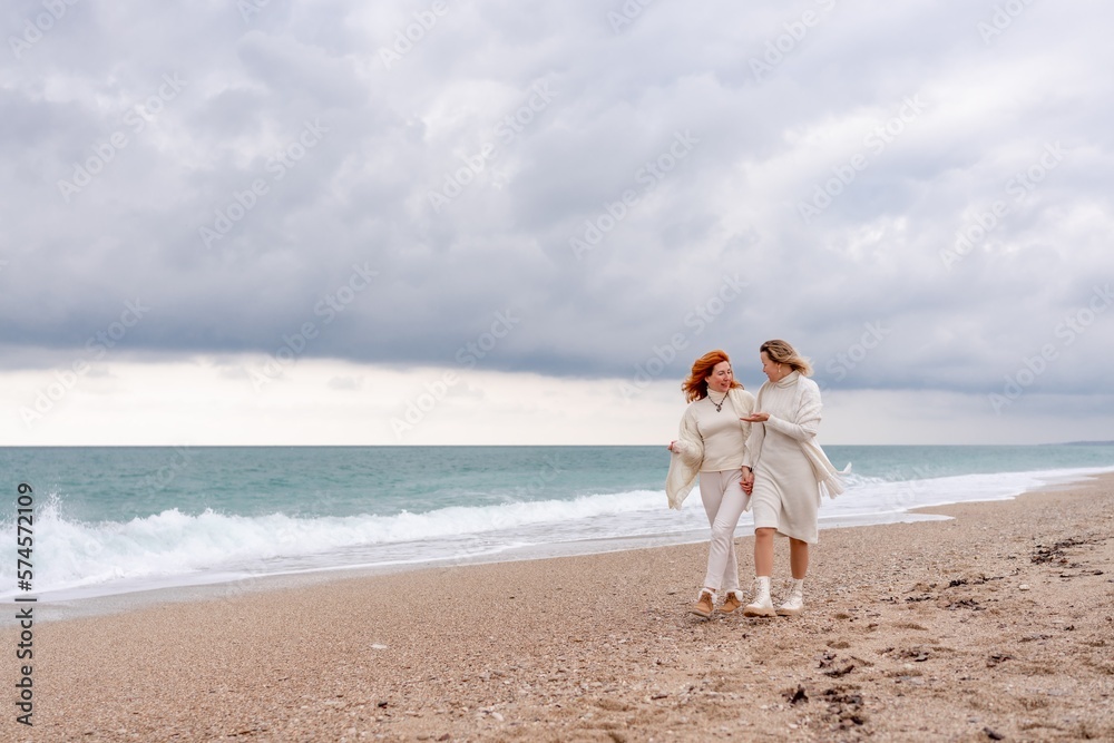 Women sea walk friendship spring. Two girlfriends, redhead and blonde, middle-aged walk along the sandy beach of the sea, dressed in white clothes. Against the backdrop of a cloudy sky and the winter Women sea walk friendship spring. Two girlfriends, redhead and blonde, middle-aged walk along the sandy beach of the sea, dressed in white clothes. Against the backdrop of a cloudy sky and the winter