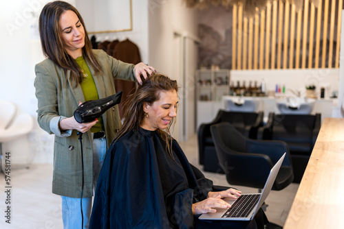 Side view of smiling hairdresser using hair dryer while female client browsing netbook and working online in beauty salon