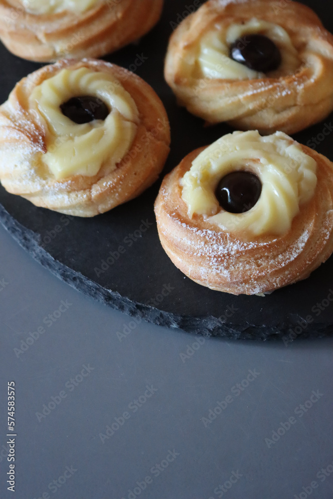 Italian traditional fried Zeppole for St. Joseph (Father’s day). Homemade sweet pastry with custard cream and black cherries on black slate stone tray on gray table