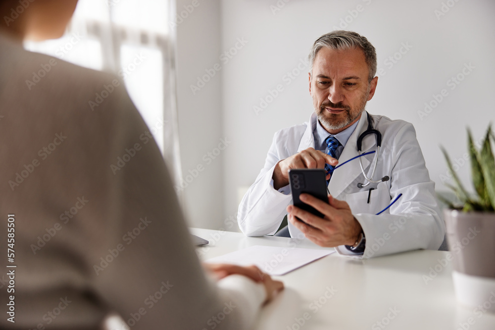 A serious doctor using a mobile phone, sitting at the office with a patient.