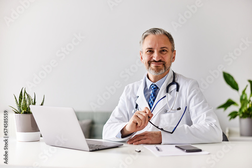 Portrait of a smiling male doctor, holding glasses and sitting in front of the laptop at the office.