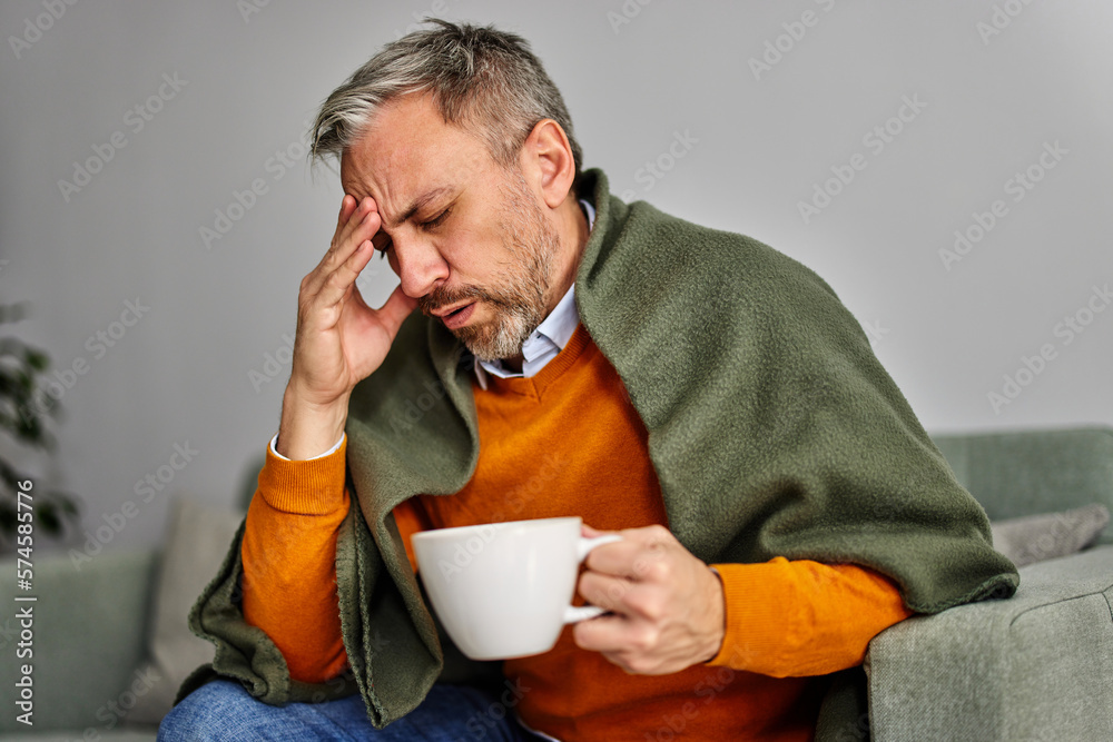 A  man holding a cup of tea, having a headache, sitting on the couch at home.