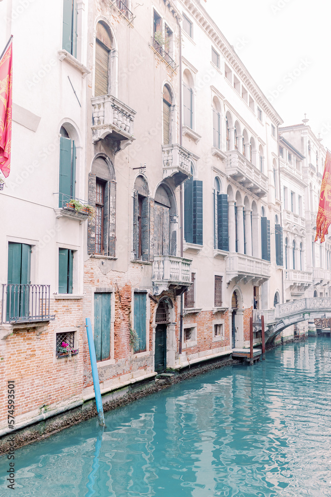 A canal runs through the canyons of houses in Venice, a beautiful bridge connects the sides. The water glitters turquoise and the sun is reflected in the brick facades of the houses.