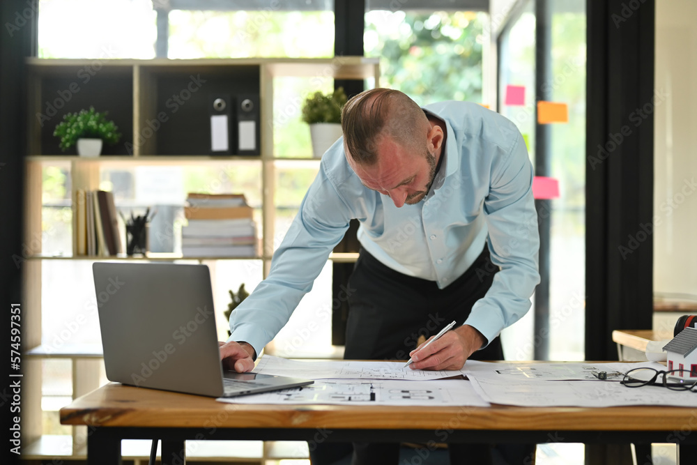 Focused engineer man in shirt using laptop and working with blueprint, examining plans at his workstation Focused engineer man in shirt using laptop and working with blueprint, examining plans at his workstation