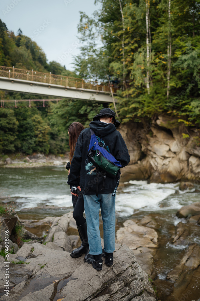 Couple of stylish tourists, man and woman, are standing on a stone near a mountain river during a hike. Vertical