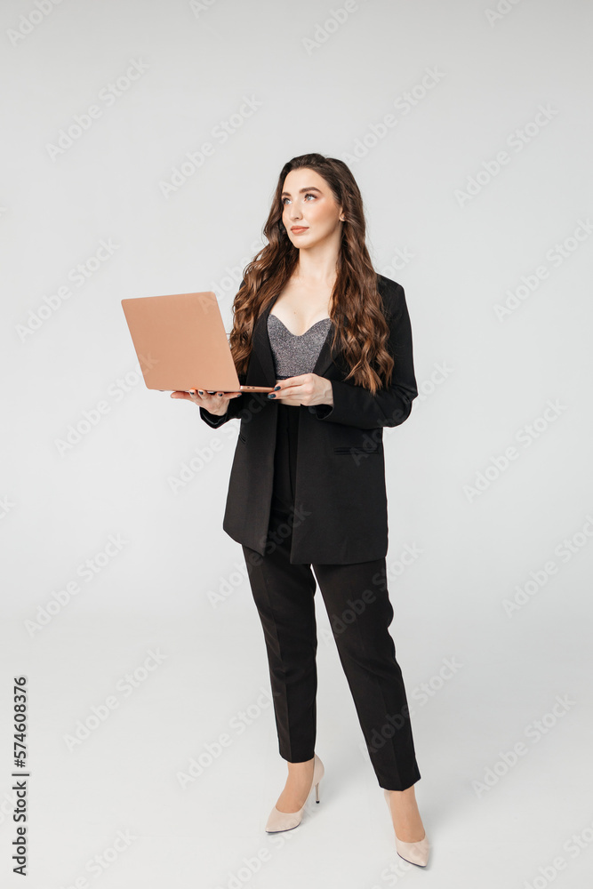 Curly adult business lady in elegant black suit standing while working on laptop against gray background Curly adult business lady in elegant black suit standing while working on laptop against gray background