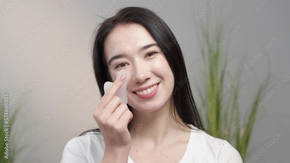 Anti-aging massage. Female beauty. Skin treatment. Smiling woman applying quartz gua sha for taking care of face posing gray background light room interior. Anti-aging massage. Female beauty. Skin treatment. Smiling woman applying quartz gua sha for taking care of face posing gray background light room interior.