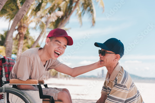 Young man with disability on wheelchair and father or volunteer or caregiver at happy time on the beach with Family Caregiving, Travel and vacation in summer, Positive photos, mental health concept.