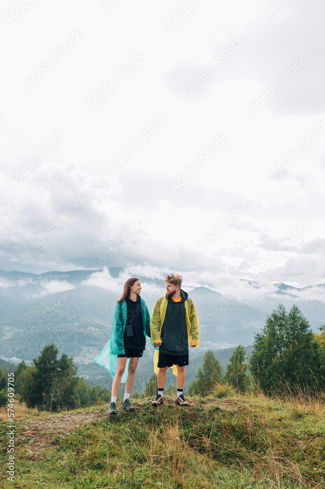 A beautiful couple man and woman in casual clothes and raincoats in rainy weather are standing in the mountains on the top against the background of a cloudy beautiful landscape.