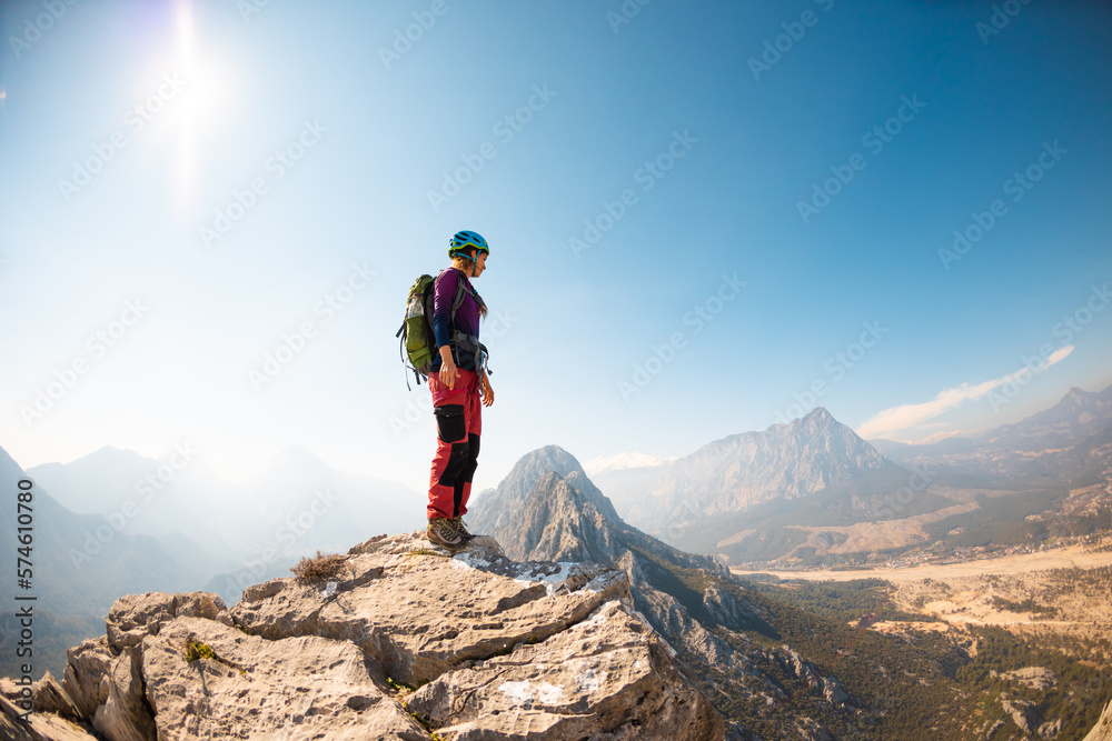 girl climber in a helmet and with a backpack stands on a mountain range against the backdrop of mountains and the sky. mountain climbing