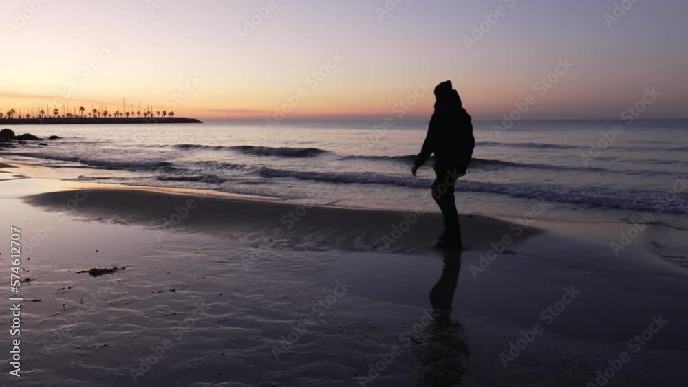 Silhouette of man walking on beach during sunrise, he stops takes photos and walks on as calm waves come ashore Silhouette of man walking on beach during sunrise, he stops takes photos and walks on as calm waves come ashore