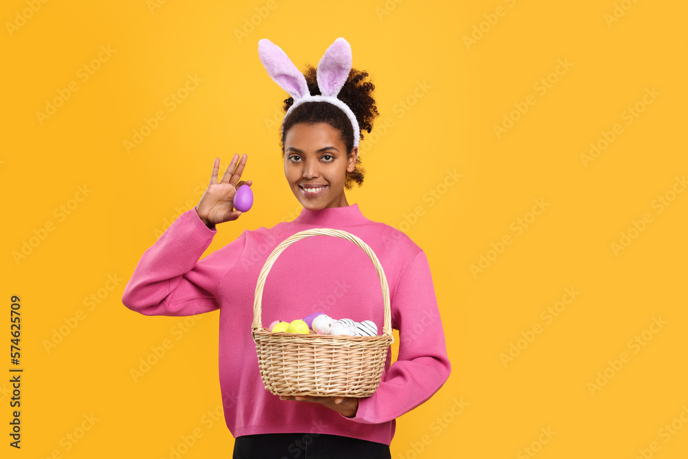 Happy African American woman in bunny ears headband holding wicker basket with Easter eggs on orange background. Space for text