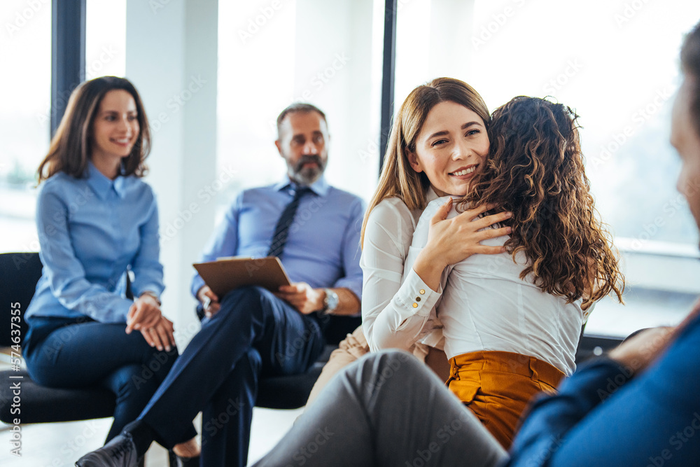 Caring female counselor hugs a female patient during a group therapy session. A young mixed-race woman hugs a mature adult woman. Caring female counselor hugs a female patient during a group therapy session. A young mixed-race woman hugs a mature adult woman.