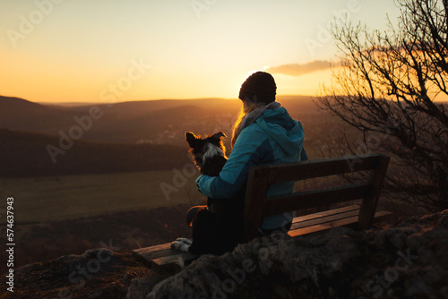 Fototapeta Naklejka Na Ścianę i Meble -  border collie puppy dog and young woman owner sitting on a bench on a mountain top at sunset seen from behind